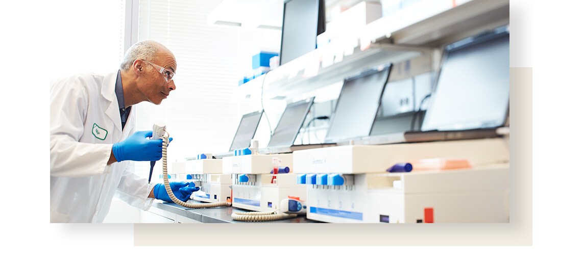 Male scientist wearing protective glasses and glue gloves is conducting a test in a lab, with line of machines in front of him.