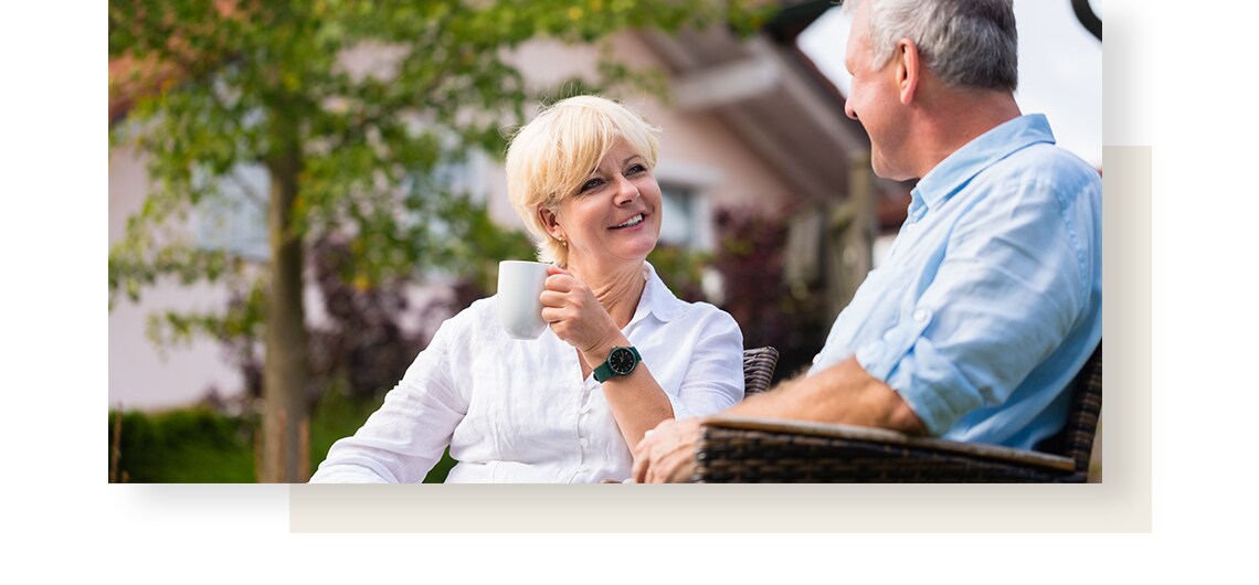 A blond woman sitting in a garden with a mug in her hand and talking to a men sitting next to her.