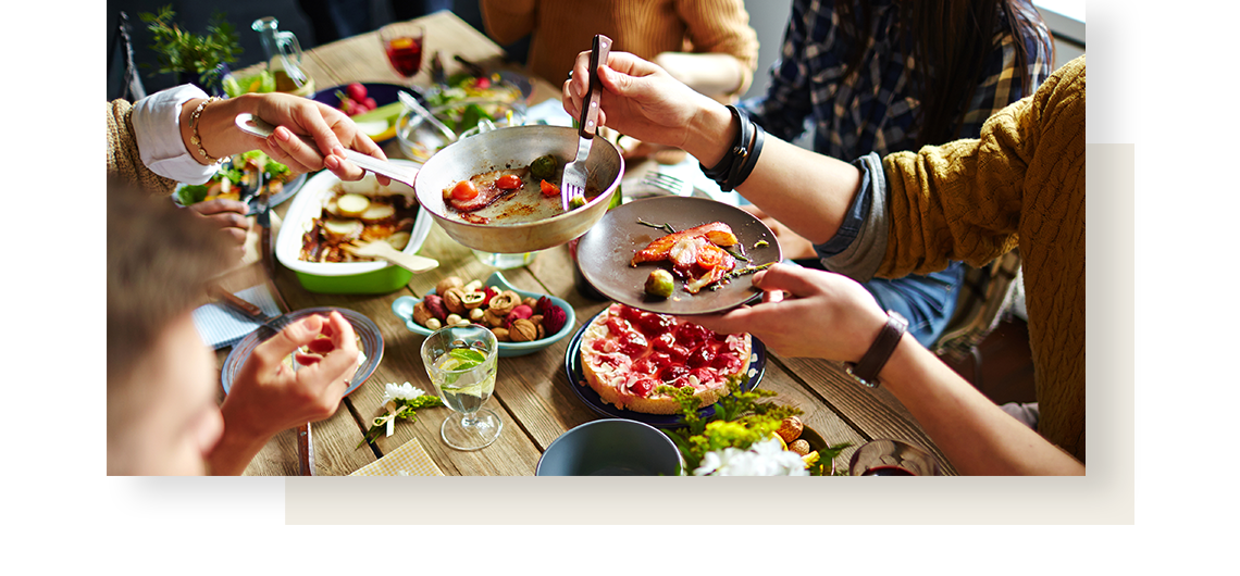 Close up of a table full of deliciously looking foods, with people sitting around it and hands handing over plates.