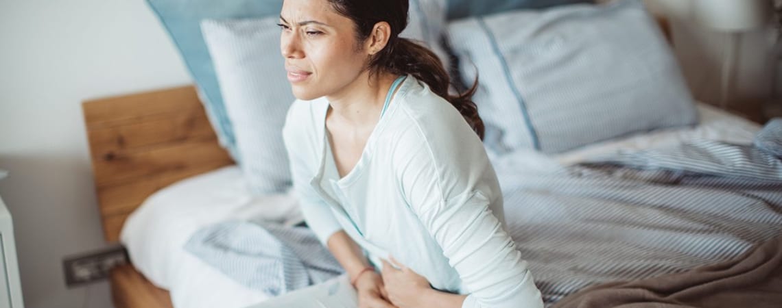 A dark-hair woman on a side of her bed, suffering from stomach pressure.