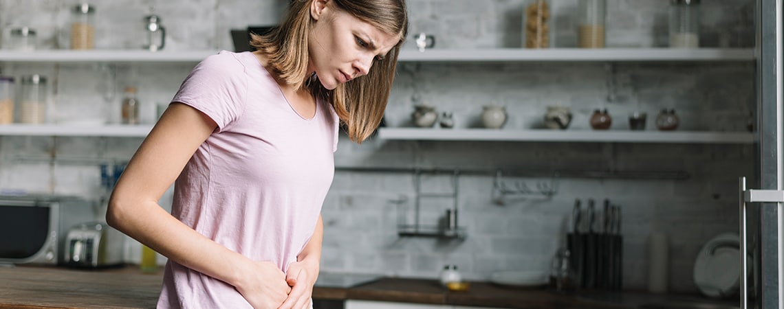 Woman suffering from irritable bowel syndrome sits on a bed, holding her stomach in pain.