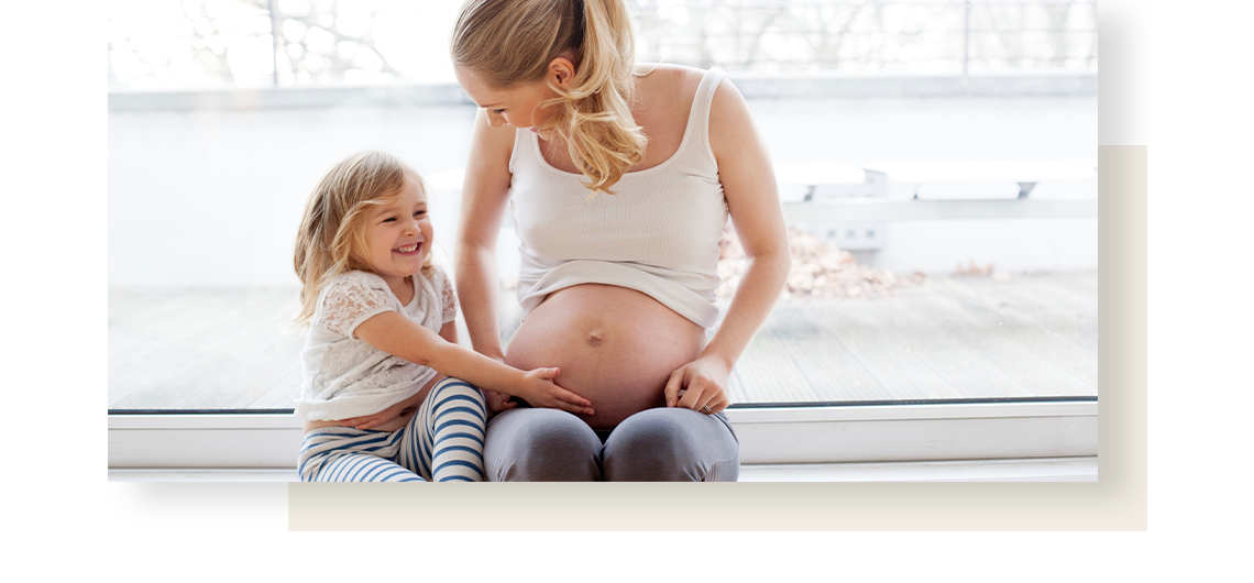 Pregnant woman sits next to her child daughter, who holds her time hand on mothers belly.