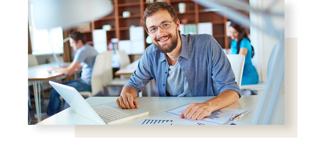 Smiling, bearded young man wearing glasses is working on his computer, with papers lying on his white desk.