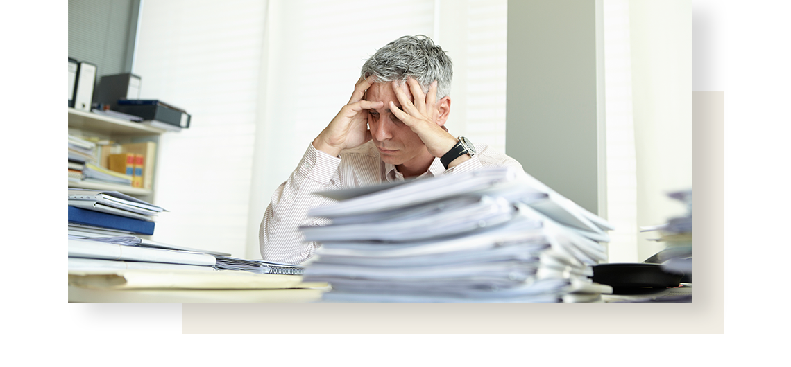 Looking stressed, gray-haired man holds his head in his hands when sitting over a pile of documents.